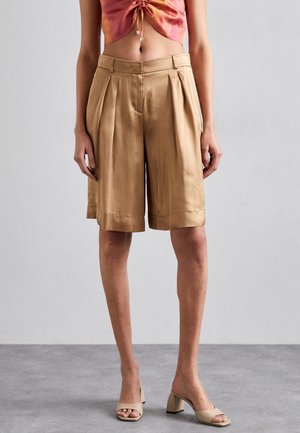 Pantaloni corti beige plissettati con una texture morbida e gambe larghe, dotati di una vita a mezz'altezza. Abbinati a sandali a tacco largo di colore chiaro.