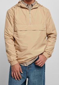 Giacca anorak beige leggera con tasca frontale, chiusura con mezza cerniera e cappuccio regolabile; indossata sopra una camicia a quadri, vestibilità comoda.