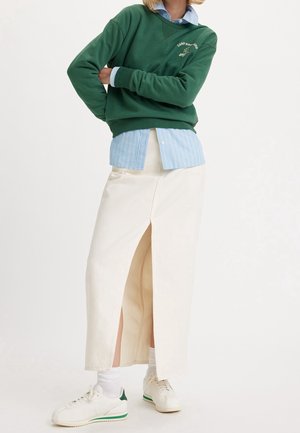 Person iført grøn sweatshirt over blå stribet skjorte, hvid midi nederdel med frontslids, hvide sokker og hvide sneakers med grønne detaljer.