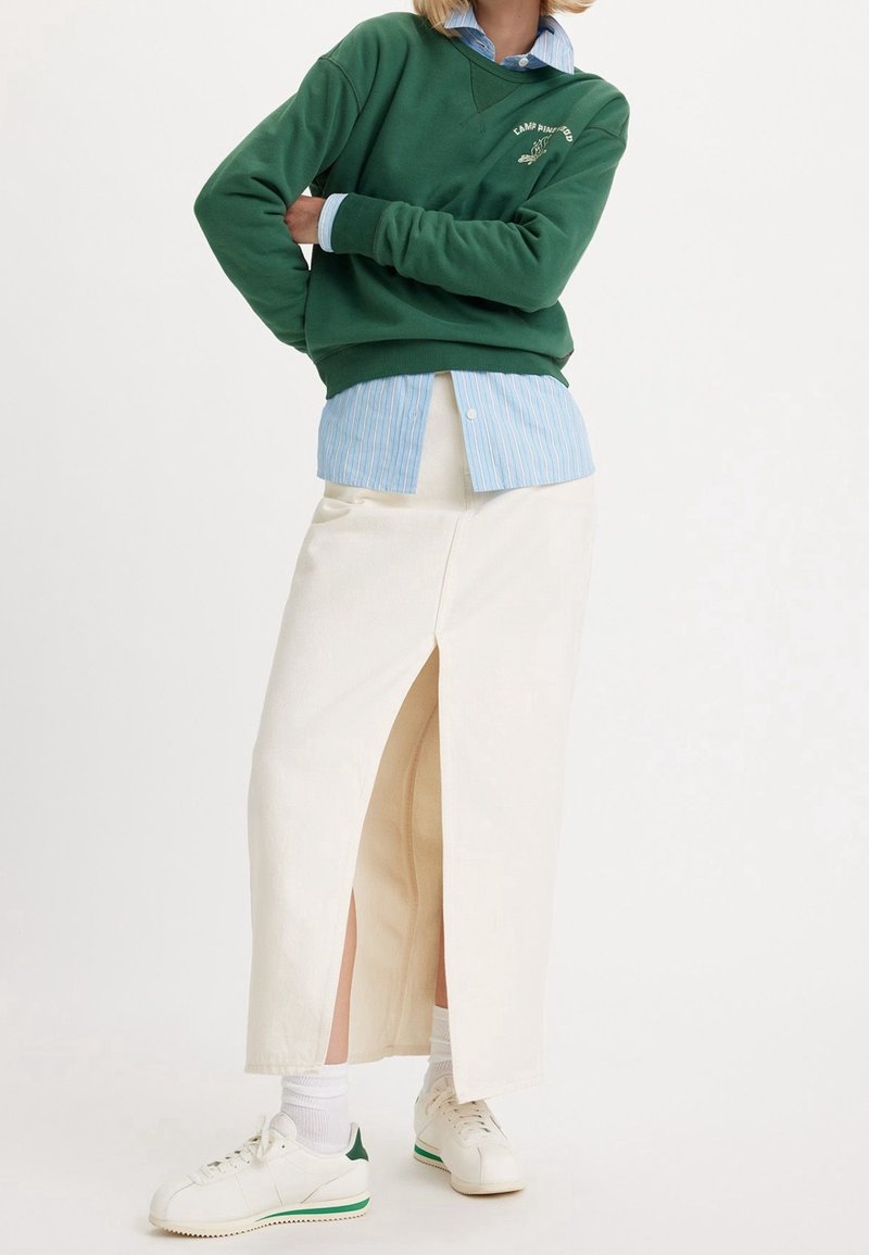 Personne portant un sweat-shirt vert sur une chemise rayée bleue, une jupe midi blanche avec une fente devant, des chaussettes blanches et des baskets blanches avec des accents verts.