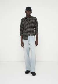 Μαύρο και πράσινο καρό πουκάμισο με κουμπιά, ανοιχτό μπλε ευρύ παντελόνι, μαύρο καπέλο και μαύρα παπούτσια με λεία υφή.