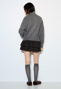 Person von hinten, trägt einen grauen, gerippten Rollkragenpullover, einen schwarz karrierten, geschichteten Minirock, kniehohe graue Socken und braune, mit Fell gefütterte Hausschuhe.