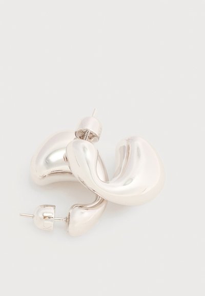 Χοντρά, γυαλιστερά ασημένια σκουλαρίκια κρίκοι με λείο, κυρτό και ελαφρώς στριφτό σχέδιο και κλεισίματα πεταλούδας στο πίσω μέρος, σε απλό φόντο.