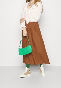 Donna che indossa una blusa bianca con volant, una gonna midi marrone, calze verdi e sneakers alte beige, mentre tiene una piccola borsa verde su sfondo bianco.