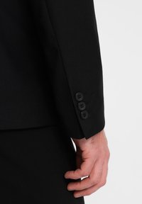 Mustat räätälöidyt pikkutakki hihat, joissa on neljä mustaa nappia, sileä kangas, istuva muotoilu, käsi näkyvissä sivussa.