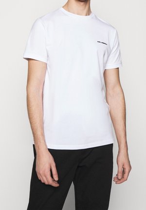 Mann trägt ein schlichtes weißes T-Shirt mit kurzen Ärmeln und Rundhalsausschnitt, kleinem schwarzem Logo auf der linken Brust und schwarze Hose.