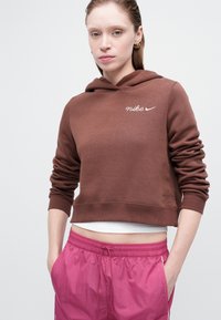 Smeđi skraćeni dukserica s kapuljačom i bijelim Nike logom, uparen s roza šortsevima na vezanje. Glatka tkanina, opušteni kroj.