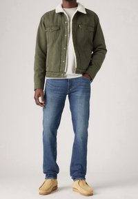 Olive green denim jacket with a cream faux shearling collar, paired with blue jeans and beige loafers. Textured fabric and standard fit.
