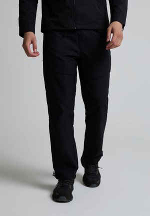 Persona che indossa pantaloni cargo neri e scarpe da ginnastica nere, in piedi davanti a uno sfondo grigio chiaro uniforme.