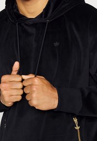 Svart zip-hoodie gjord av mjuk sammet. Har en liten svart logotyp på bröstet, justerbara dragsnören och sidofickor med guldblixtlås.