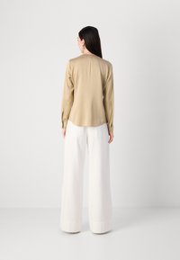 Camicia lunga beige con una texture liscia, caratterizzata da una cucitura posteriore. Abbinata a pantaloni bianchi larghi, con tasche visibili sul retro.