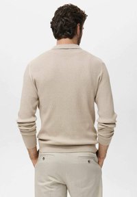 Suéter beige de canalé con cuello redondo, mangas largas y un diseño ajustado; combinado con pantalones de color claro que presentan detalles en los bolsillos traseros.