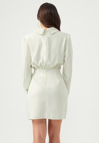 Femme aux longs cheveux bruns portant une mini-robe ajustée à manches longues de couleur crème avec une fermeture éclair au dos et une taille froncée, vue de dos.