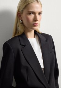 Blazer nero con revers appuntiti, tasca singola, indossato sopra un top bianco a coste. Capelli lunghi e lisci biondi, trucco minimale, orecchini in argento.