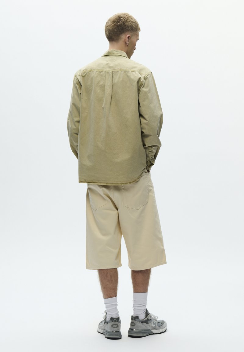 Chemise boutonnée couleur olive clair à manches longues, assortie à un short beige arrivant aux genoux. Chaussures de sport grises, chaussettes blanches, et fond neutre.