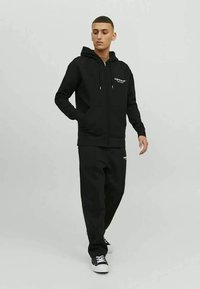 Ung mand iført sort zip-up hættetrøje og matchende sorte sweatpants med sneakers, stående foran en ensfarvet lys grå baggrund.