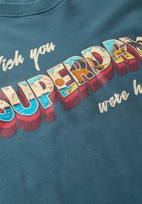 Sudadera verde azulado con un gran texto multicolor de diseño retro que dice "Ojalá estuvieras aquí" y "SUPERDA". Textura suave.