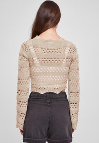 Beige strikket crop genser med lange ermer og åpningsmønstre, bølget nederkant og teksturert garn. Kombinert med mørke shorts.