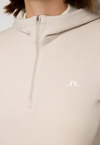 Felpa con zip beige dal tessuto morbido, cappuccio regolabile e logo bianco sul petto. La cerniera è centrata e discretamente progettata.