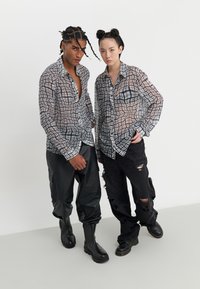 To personer iført gennemsigtige, ternede skjorter med knapper i nuancer af sort og hvid. Den ene har sorte læderbukser og støvler på, den anden har slidte jeans.