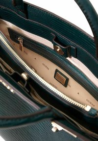 Zielona torebka o fakturowanej powierzchni z złotymi elementami, zamkiem błyskawicznym oraz wnętrzem wyściełanym beżową tkaniną z powtarzającym się wzorem logo „GUESS”.