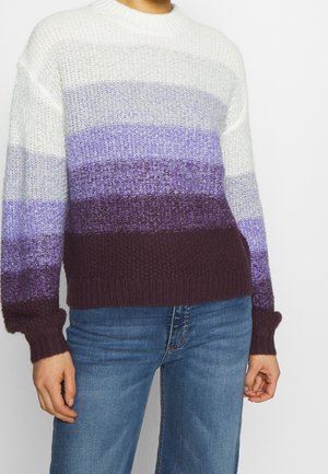 Personne portant un pull en maille avec des bandes horizontales de couleur blanc, violet clair, violet moyen et violet foncé, associé à un jean bleu.