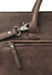 Detail van een bruine leren handtas met een ritsvak, metalen hardware en een karabijnhaakbevestiging op een gestructureerd oppervlak.