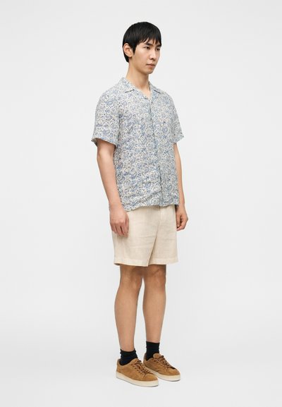 Chemise à manches courtes à motif floral bleu et crème, associée à un short en lin beige et des baskets en daim brun.