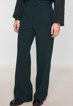 Pantalons larges en tissu vert foncé, avec une texture lisse, une taille ajustée et des poches latérales, associés à des chaussures noires.