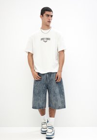Hvid t-shirt med "Guns N' Roses" grafik, parret med blå denim shorts med et falmet stribemønster og hvide sneakers med blå accenter.
