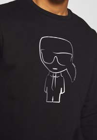 Czarna bluza z kapturem z srebrnym nadrukiem stylizowanej postaci w okularach przeciwsłonecznych, w garniturze i krawacie, na przodzie.