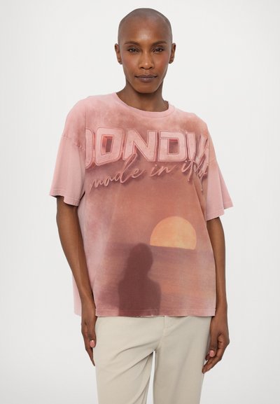 T-shirt en coton surdimensionné de couleur rose avec un imprimé graphique d'un soleil se couchant sur l'eau, avec du texte blanc en gras et une finition douce et texturée.