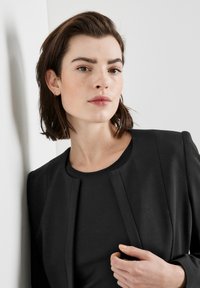 Schwarzer maßgeschneiderter Blazer mit rundem Ausschnitt und schlankem Design. Glatte Textur, ohne sichtbare Verschlüsse und mit minimalistischem Styling.