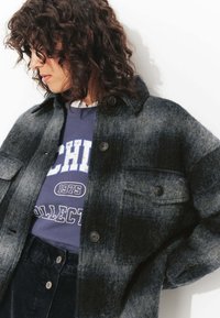 Veste en laine grise et noire ombragée avec deux poches poitrine, fermeture à boutons et coupe décontractée, portée sur un sweat-shirt graphique bleu.