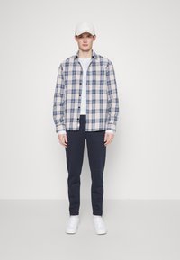 Camicia a quadri in crema, blu e navy, indossata sopra una maglietta bianca. Pantaloni slim navy e sneakers bianche completano il look.