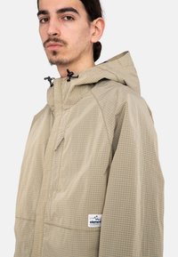 Chaqueta ligera beige con un patrón de rejilla, que cuenta con capucha, cordones ajustables y un pequeño parche de logo en el lado izquierdo.