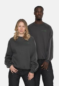 Sudaderas grandes de color gris oscuro con pu�ños y bajos acanalados. Presenta un logo verde en el pecho y acentos de rayas en los lados. Combinadas con jeans negros.
