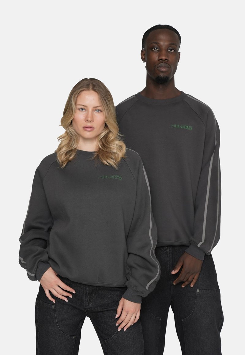 Sudaderas grandes de color gris oscuro con puños y bajos acanalados. Presenta un logo verde en el pecho y acentos de rayas en los lados. Combinadas con jeans negros.