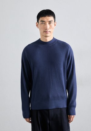 Marineblauer Rippstrickpullover mit Rundhalsausschnitt und langen Ärmeln, der ein strukturiertes Muster und eine lockere Silhouette aufweist, kombiniert mit schwarzen Hosen.