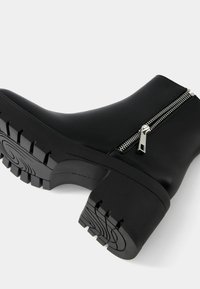 Bottine noire en matériau lisse, avec une semelle en caoutchouc épaisse à profil texturé et un détail de fermeture éclair métallique sur le côté.