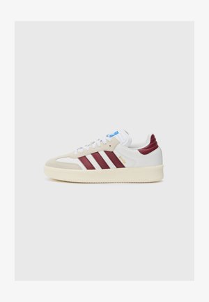 Zapatilla blanca con rayas burdeos, puntera de ante beige y suela texturizada en blanco roto. Presenta una etiqueta azul en la lengüeta y letras doradas.