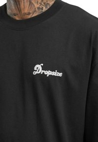 Must musta T-särk, millel on vasakul rinnal valge "Dropsize" logo tikandina. Ümar kaelus, lühikesed varrukad, sile tekstuur.