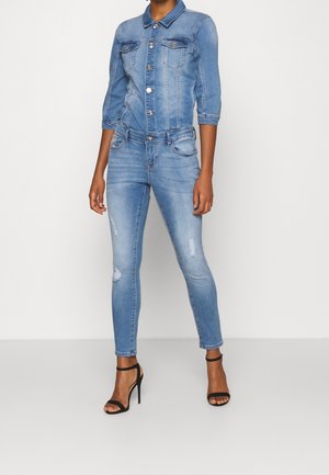 Vrouw draagt een aansluitend lichtblauw denimjack met driekwartmouwen, bijpassende distressed skinny jeans en zwarte sandalen met hoge hakken.