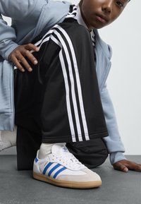 Hvite Adidas Samba-sneakers med tre blå striper, gummisåle og teksturert tåhette. Svart treningsdrakt med hvite side-striper og lyseblå hettegenser.