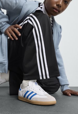 Sneaker Adidas Samba bianche con tre strisce blu, suola in gomma e punta testurizzata. Tuta nera con strisce laterali bianche e felpa leggera blu chiaro.