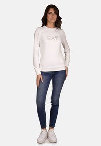 Weißer Langarm-Sweatshirt mit "EA7"-Logo aus glänzendem Material, kombiniert mit engen, dunkelblauen Jeans und weißen Sneakers. Einfaches Design.