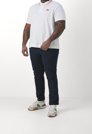 Polo shirt blanc avec col rouge, affichant un logo brodé. Assorti à un pantalon marine foncé et des baskets blanches avec des accents verts.