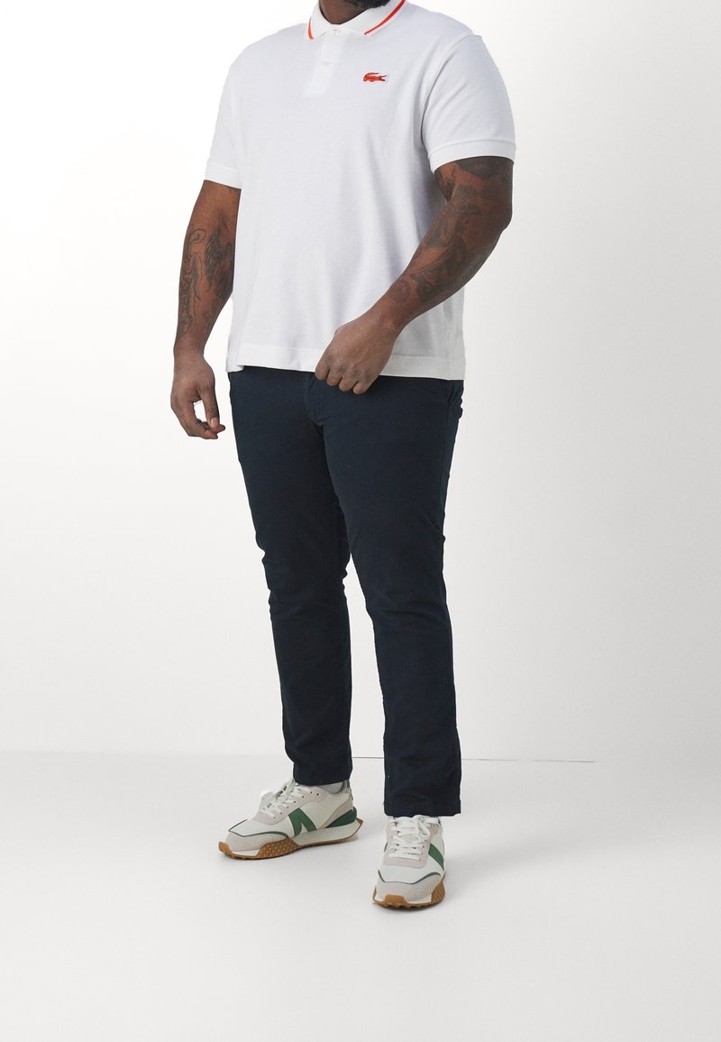 Polo shirt blanc avec col rouge, affichant un logo brodé. Assorti à un pantalon marine foncé et des baskets blanches avec des accents verts.