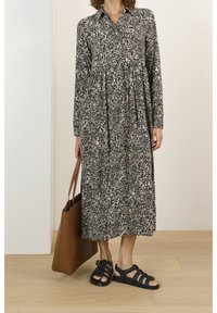Donna che indossa un vestito a stampa leopardata con maniche lunghe e lunghezza midi, mentre tiene una borsa a tracolla in pelle marrone, indossando sandali neri intrecciati, in piedi su un pavimento in legno.