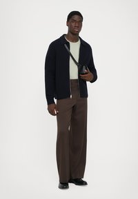 Hombre con un cárdigan azul marino con cremallera, camisa verde clara, pantalones anchos marrones, zapatos negros y sosteniendo un pequeño bolso cruzado negro.